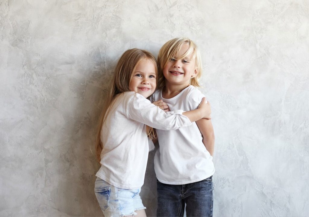 Ein Bild von zwei Kinder, die lachen und sich umarmend vor einer Wand stehen.