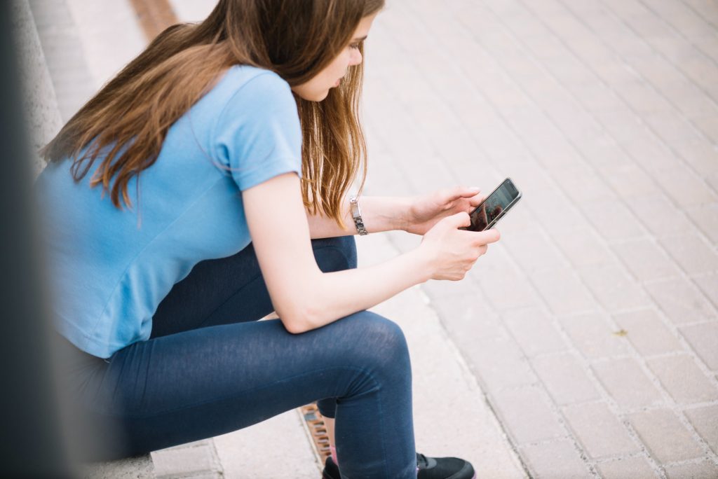 Eine Aufnahme von einer Jugendlichen die auf ein paar Stufen sitzt und an ihrem Handy ist.